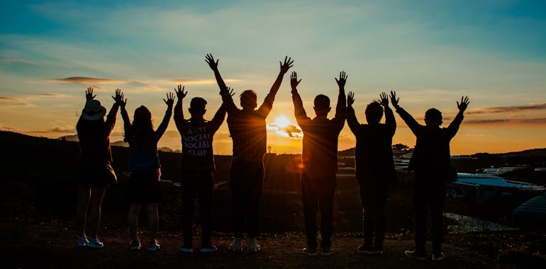 A diverse group of friends raises their arms in celebration against a vibrant sunset backdrop.
