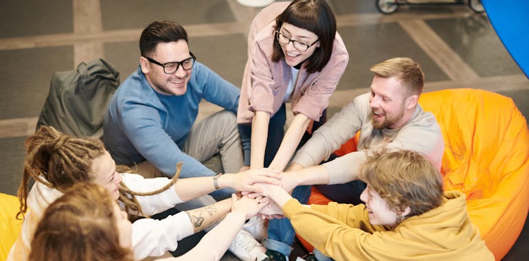 A diverse group of adults sit indoors, joyfully stacking hands together, symbolizing teamwork and camaraderie.